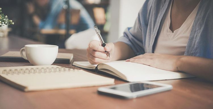 woman writing on a notebook beside teacup and tablet computer